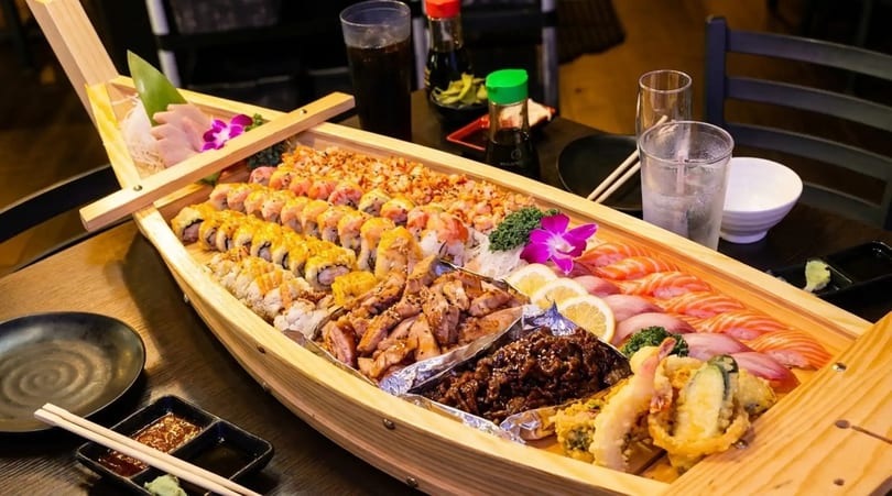 A large wooden boat filled with various sushi rolls, sashimi, grilled chicken, and tempura.